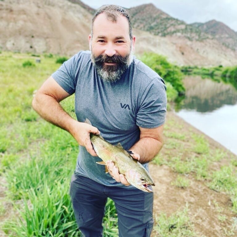 A man smiling while holding a large fish caught at a scenic outdoor location.